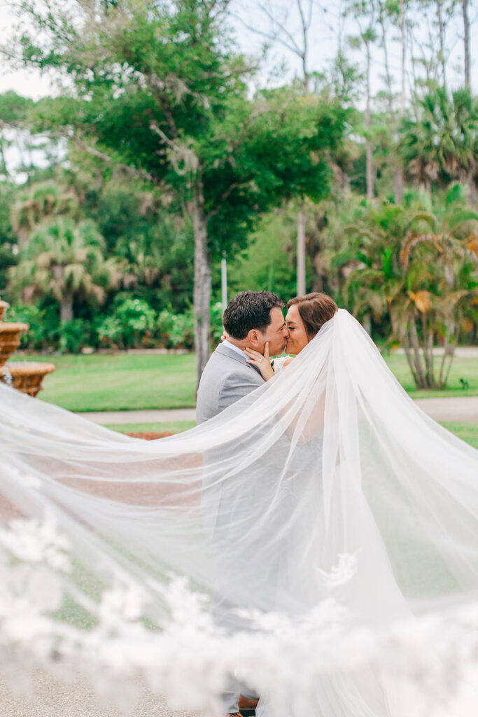 A couple sharing a kiss with the bride’s veil flying in front of the stone fountain in the courtyard at Bonita Ranch, a wedding venue in Melbourne, Florida.